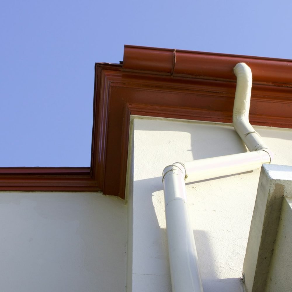 Painted fascia and gutter at a roof corner with a downspout on a stucco wall
